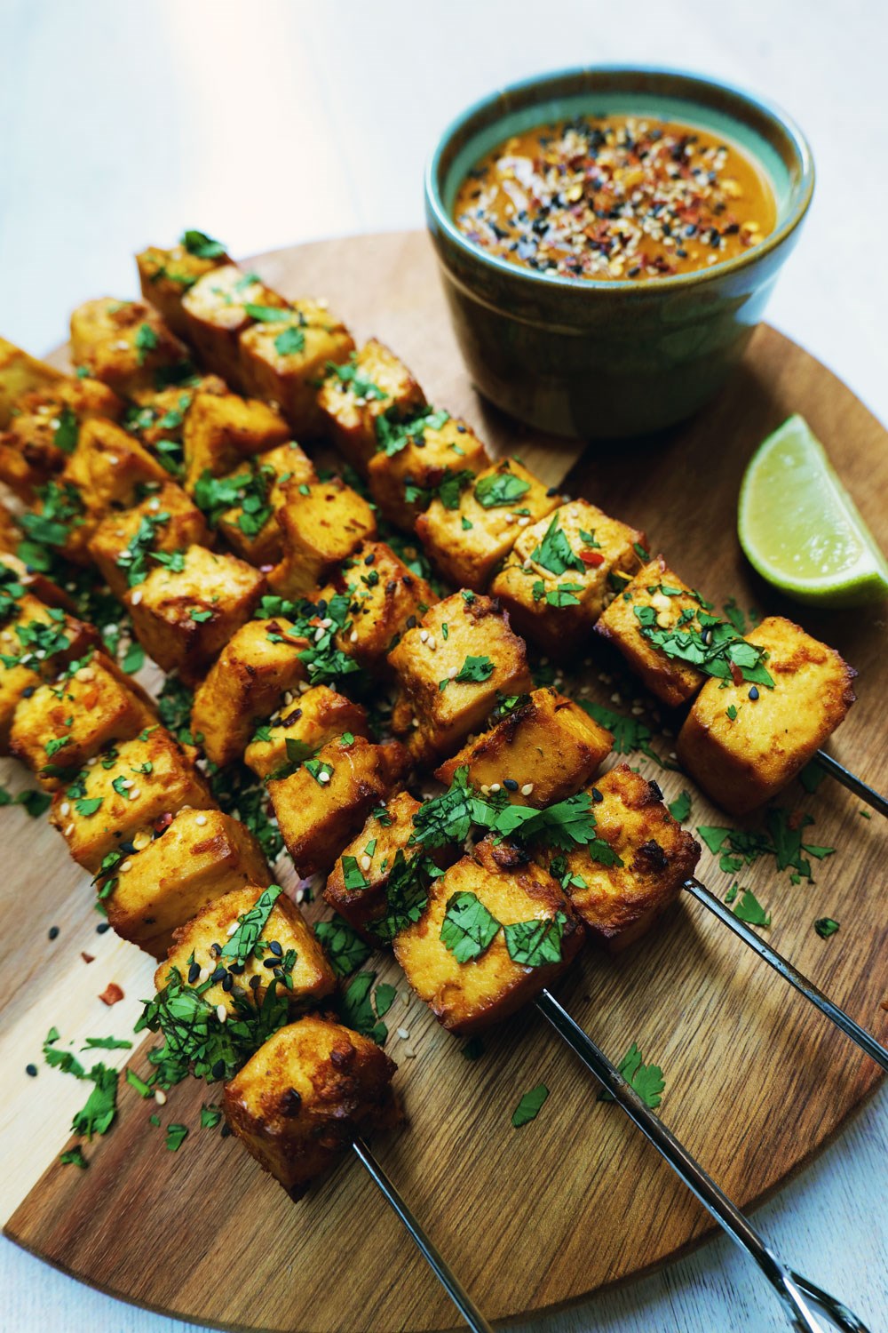 skewered tofu on a wooden chopping board with peanut sauce and garnished with herbs, sesame seeds and chillies