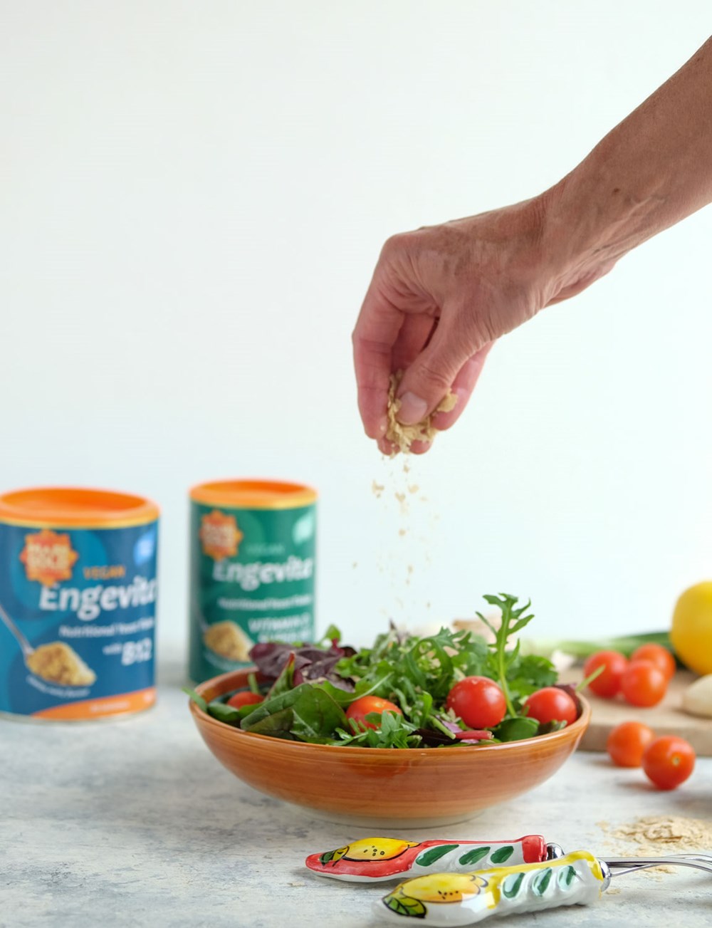 A hand sprinkling nutritional yeast flakes over a bowl of salad with Marigold products in the background