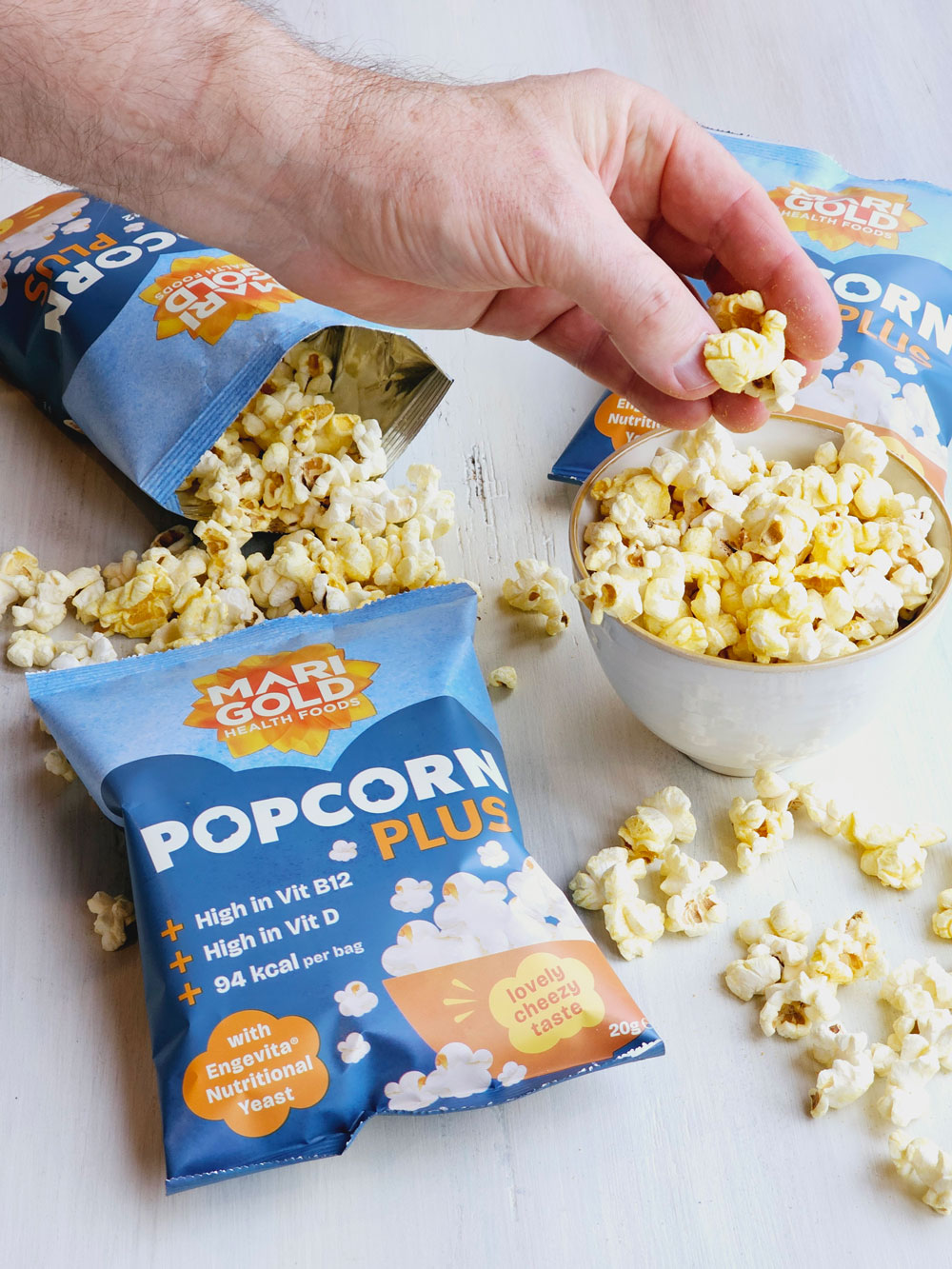 A hand reaching for a small bowl of Marigold Popcorn. Around it is packs of the popcorn and scattered popcorn.